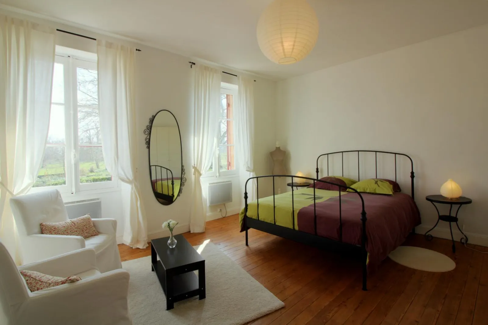 Bright and spacious bedroom featuring a black wrought iron bed, white armchairs by large windows, oval mirror, wooden flooring and minimalist décor at the Maison des Hôtesses in Bordeaux.