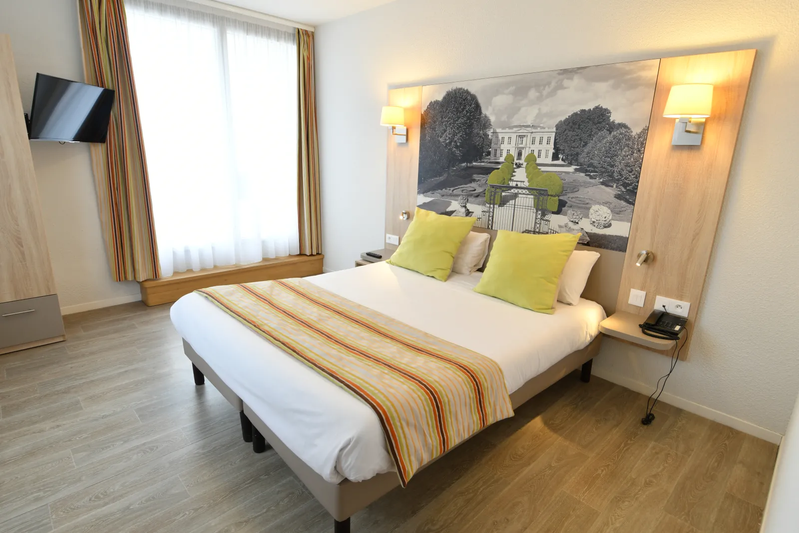 Modern hotel room in Bordeaux featuring a double bed with white linens and green cushions, large artwork of a castle above the headboard, striped curtains on the window, wall-mounted flat screen TV, and light wooden flooring.