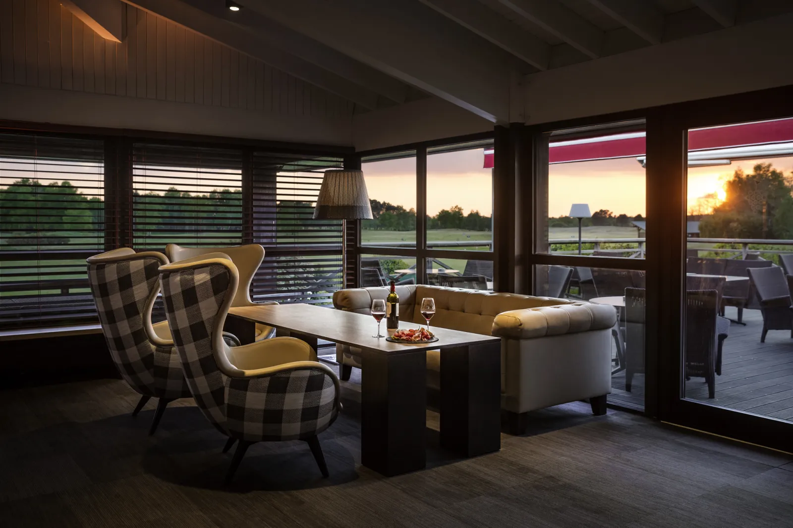 Elegant lounge at Le Club restaurant, Golf du Pian Médoc in Bordeaux, featuring checkered armchairs, beige sofa, black coffee table, panoramic windows overlooking the greens at sunset, and outdoor terrace.
