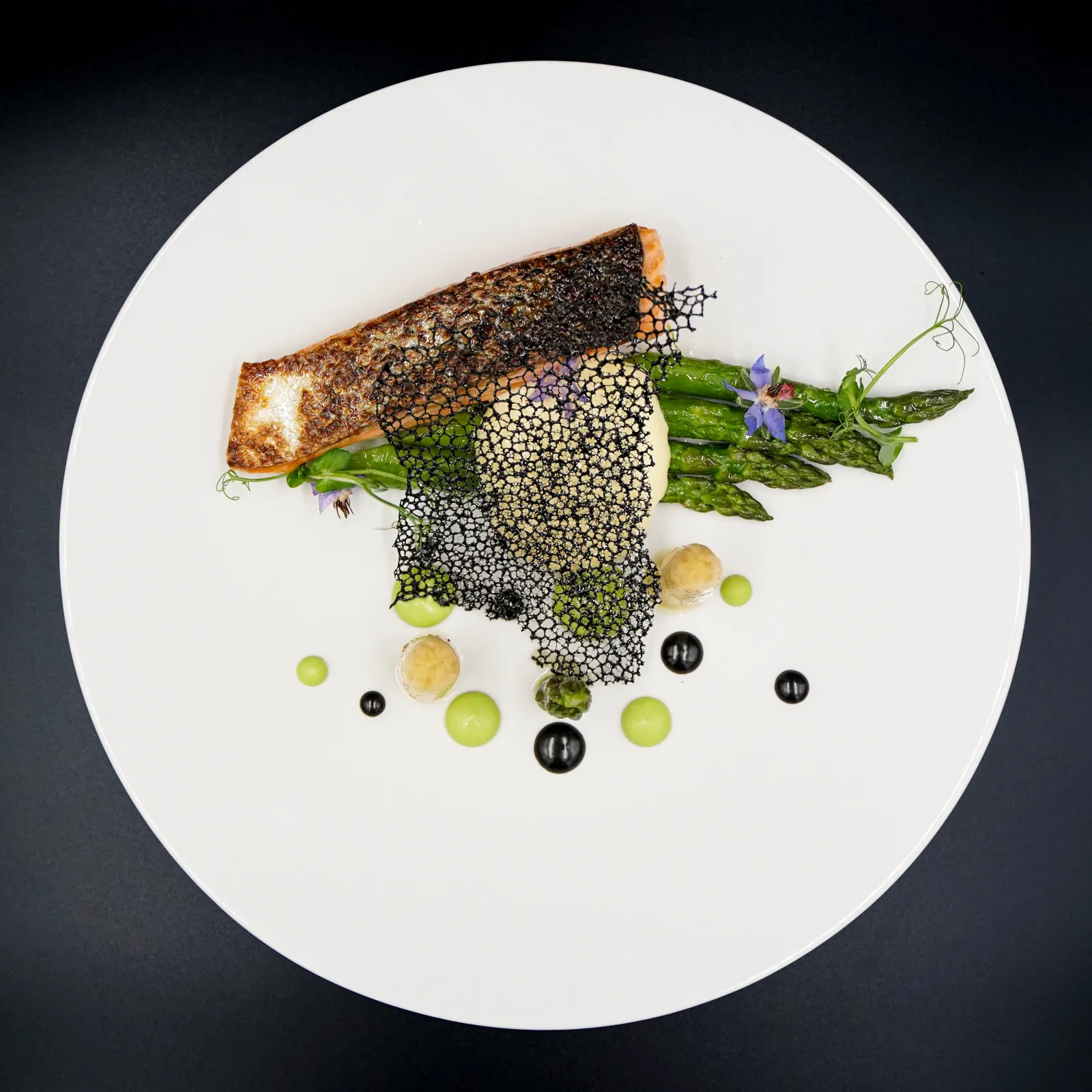 Fine dining plate featuring a golden-crusted fish fillet, garnished with green asparagus, caviar, fruit pearls, and edible flowers on a white background