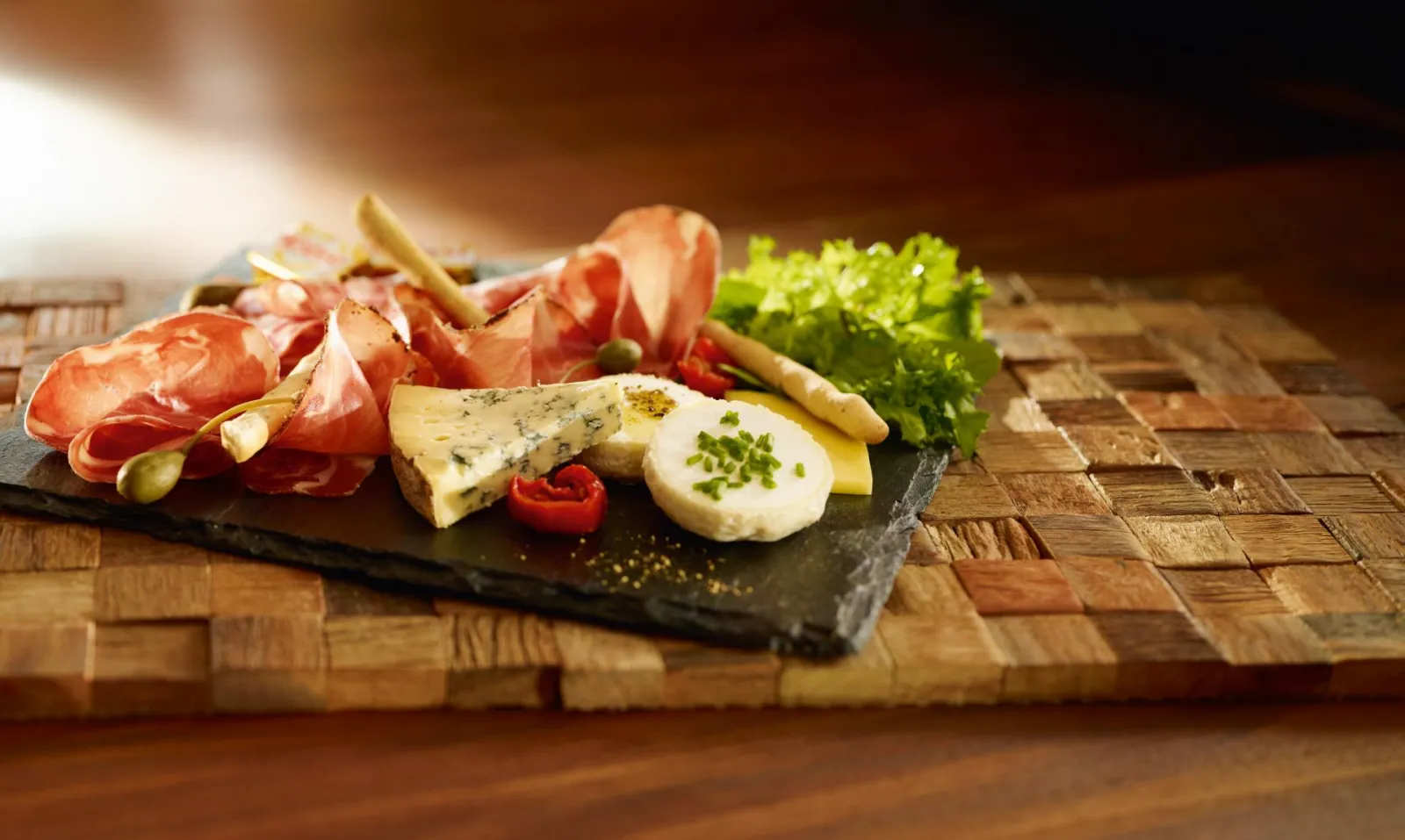 A charcuterie and cheese board on a black slate with Parma ham, blue cheese, fresh mozzarella, toasted breadsticks, pickles, lettuce and fresh herbs on a wooden board in Bordeaux.