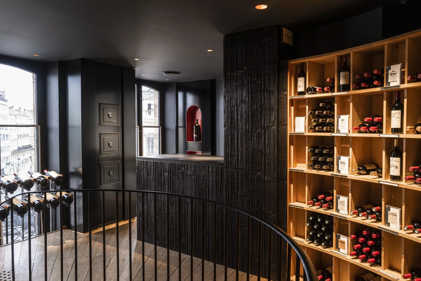 Modern wine cellar interior in Bordeaux featuring wooden shelves filled with red wine bottles, a black counter with a red wine alcove, city views through windows, and a metal railing.