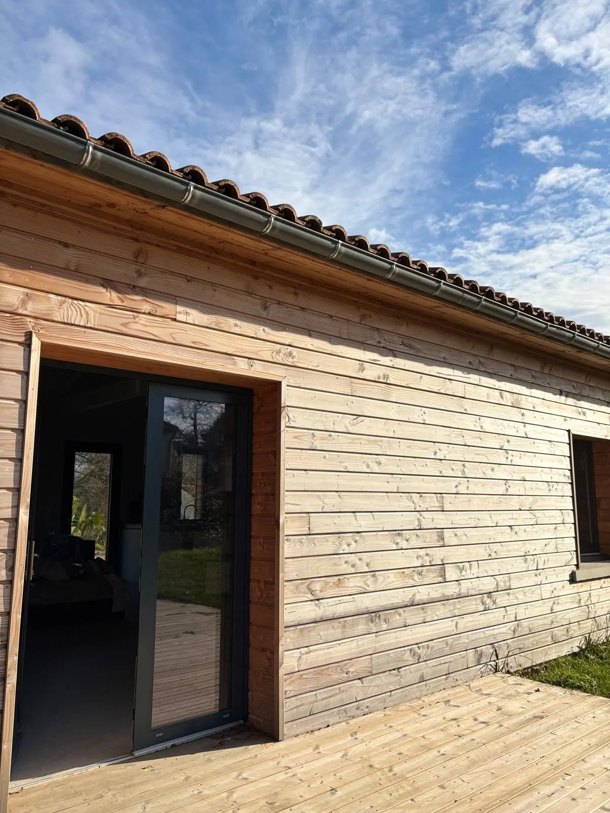 Modern wooden eco-cottage with light-colored wood cladding, black door and windows, brown tile roof, wooden terrace, set against a partly cloudy blue sky