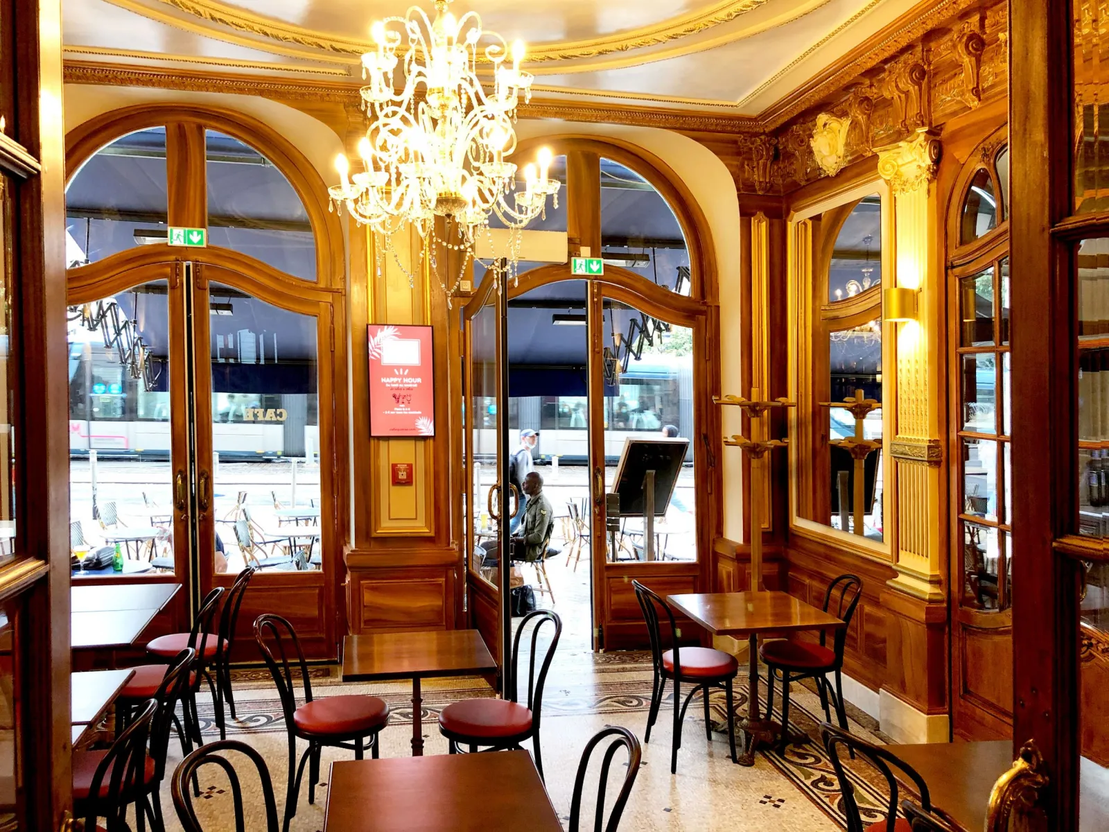 Elegant interior of QuatrèS Café in Bordeaux featuring golden wood paneling, crystal chandelier, wooden glass doors, and classic furniture with outdoor terrace visible.
