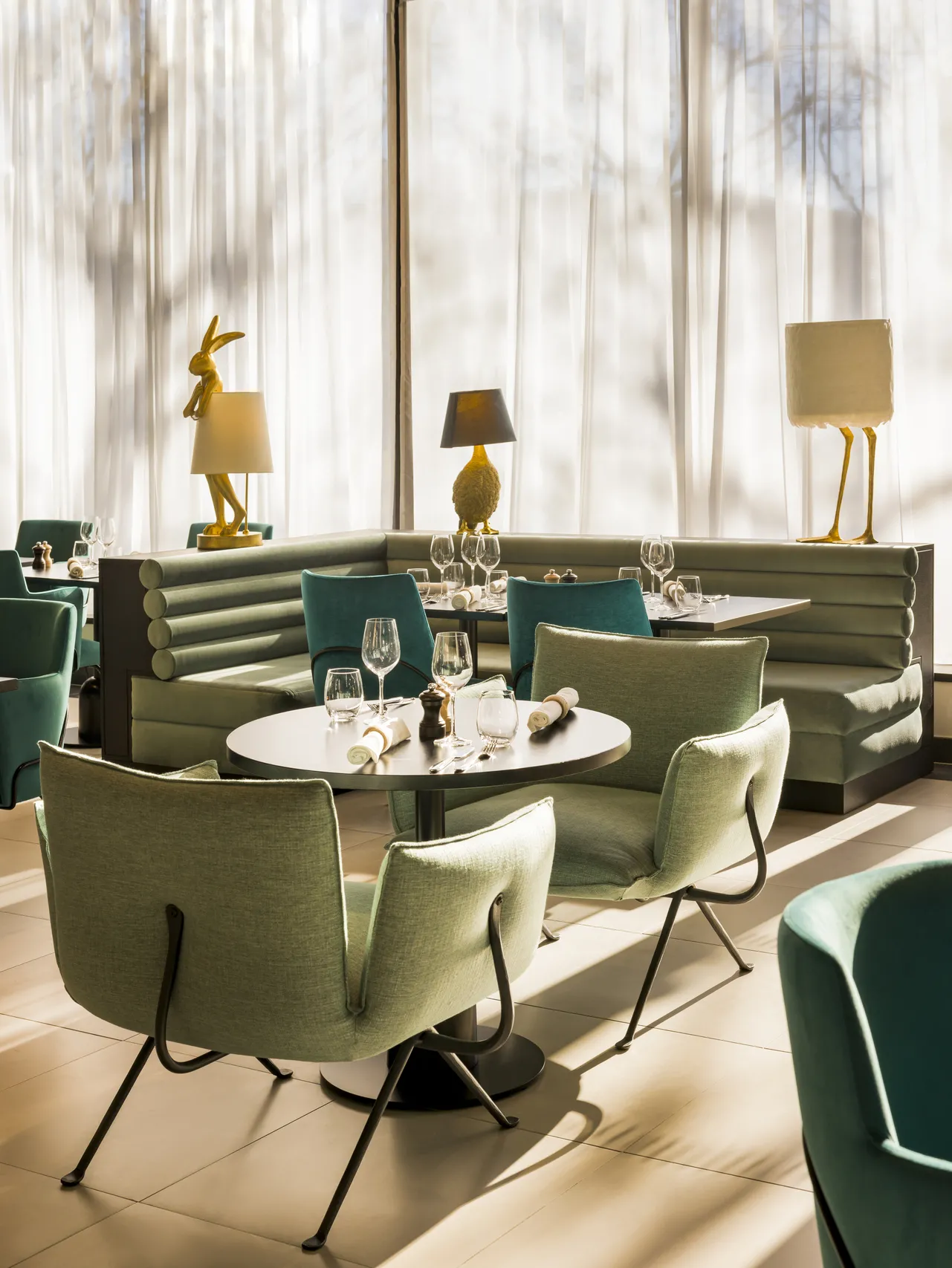Elegant dining area at Bada restaurant in Bordeaux featuring green banquette seating, designer chairs, white round table, golden geometric lamps, and bright floor-to-ceiling windows.