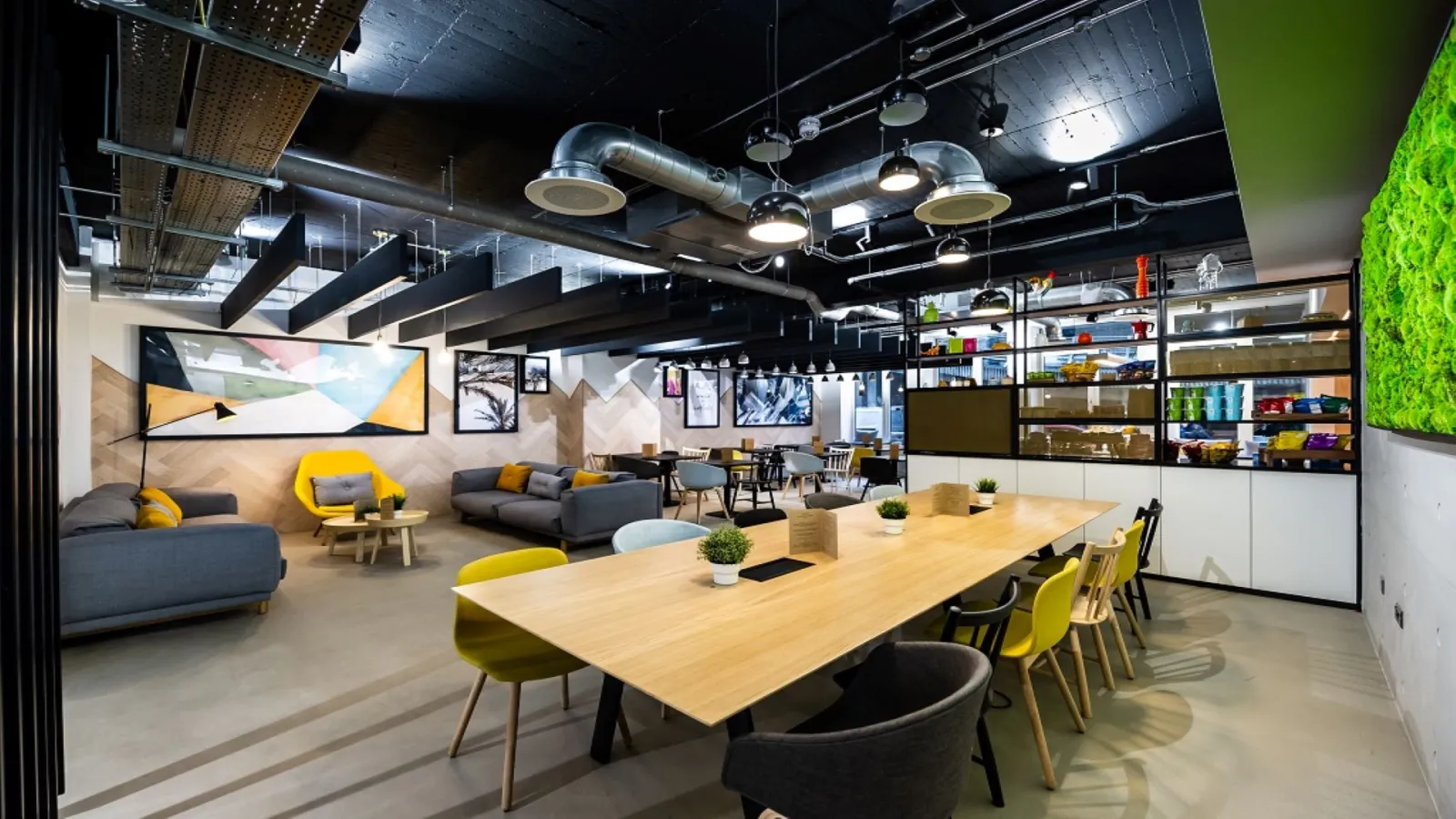Modern common area of a Bordeaux aparthotel featuring a wooden dining table in the foreground, relaxation area with gray sofas and yellow accents, wall-mounted shelving, industrial ceiling with exposed ductwork and pendant lights, living green wall on the