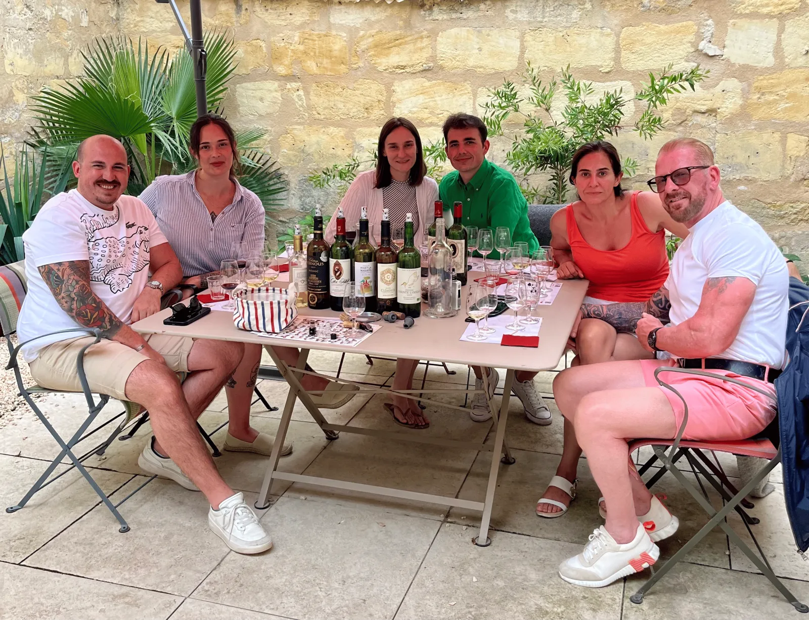 Six smiling people seated around a wine tasting table with multiple wine bottles, glasses and tasting accessories, in a paved courtyard in front of a stone wall in Bordeaux.
