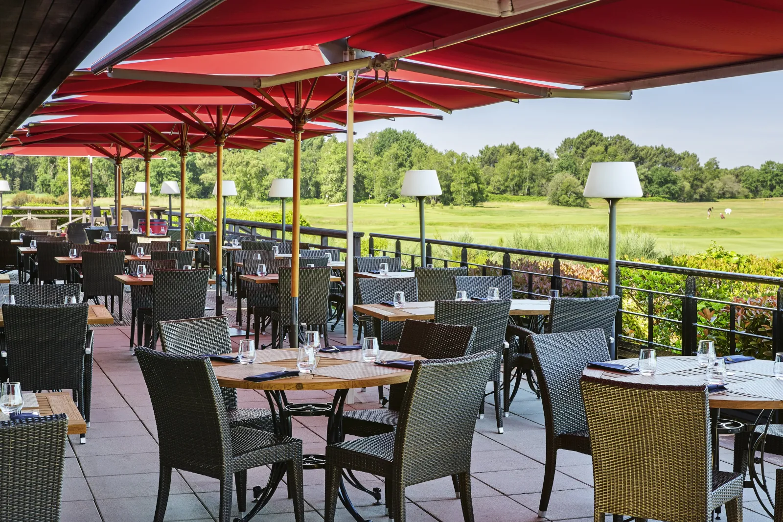 Shaded outdoor terrace of Cabot Bordeaux MGallery restaurant featuring wooden tables, gray wicker chairs, red umbrellas, and panoramic views of a green park.