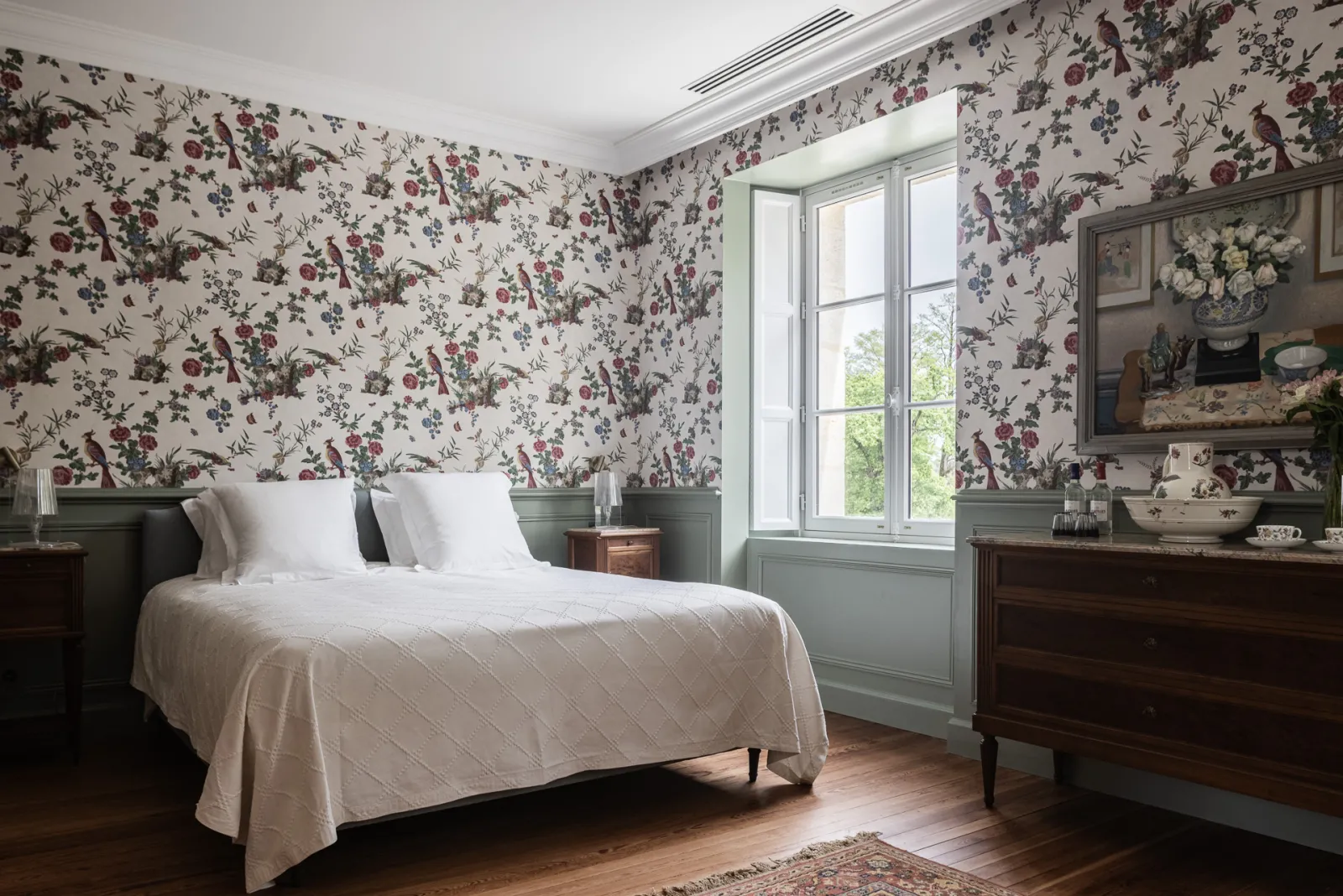 Elegant bedroom at Château Sénéjac featuring a white bed, floral wallpaper with birds and roses pattern, sage green paneling, wooden flooring, window overlooking the garden, and antique walnut furniture.
