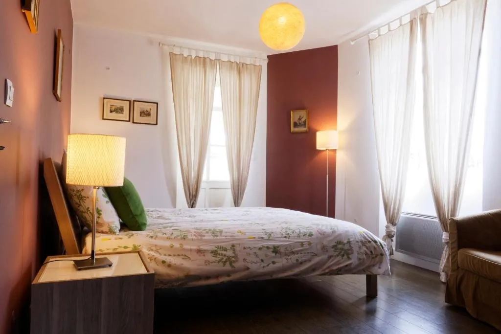 Elegant bedroom featuring a double bed, burgundy and cream walls, large windows with beige curtains, gold table lamps, yellow ceiling light, and vintage furniture at La Bastide de Siran, Bordeaux.