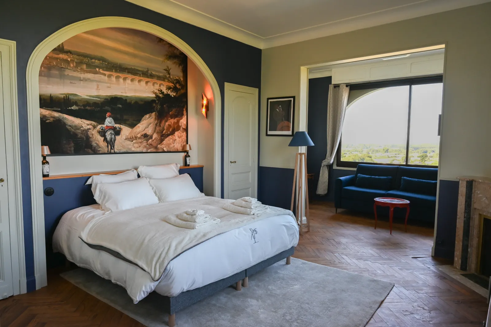 Spacious bedroom at Chalet des Perchés in Bordeaux featuring a white bed under an architectural arch with a scenic mural, navy blue and beige walls, large bay windows with panoramic views, and a blue sofa seating area.