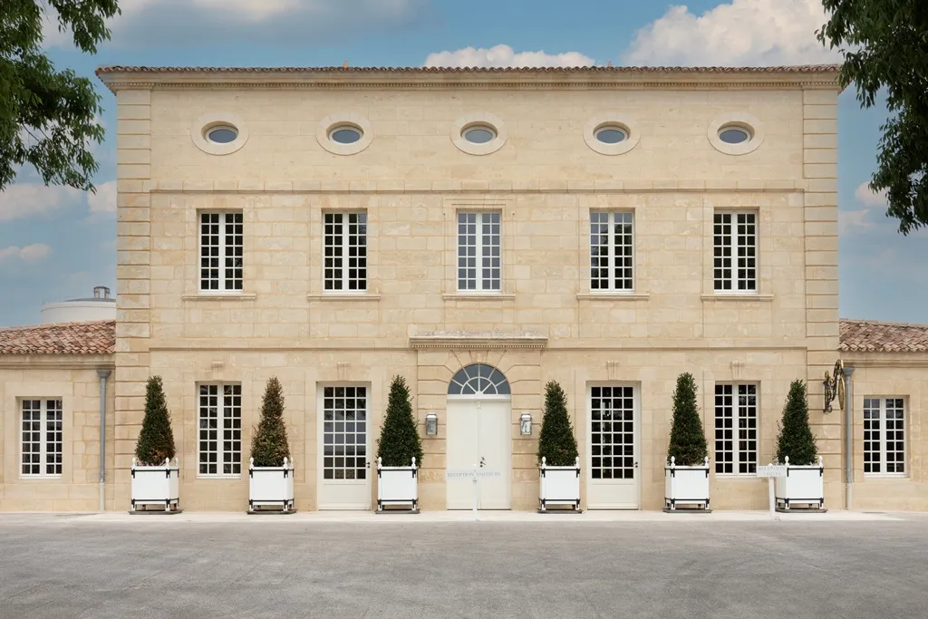 Elegant facade of the Cave du Château in Bordeaux, a stone building with five round windows above and five French doors at ground level, flanked by green cypress topiaries in white pots, under a partly cloudy sky.
