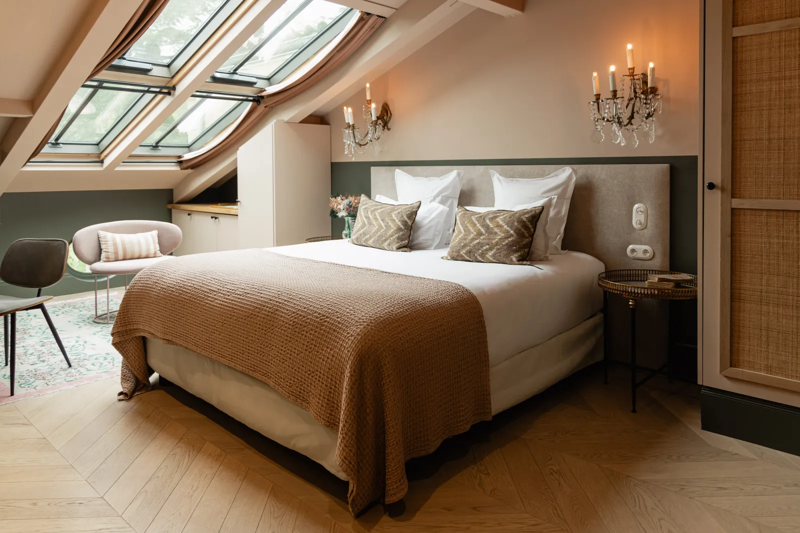 Elegant bedroom in a castle featuring a king-size bed with gray headboard, crystal chandeliers, modern chairs, skylights, exposed beams and wooden flooring - Château Léognan, Bordeaux