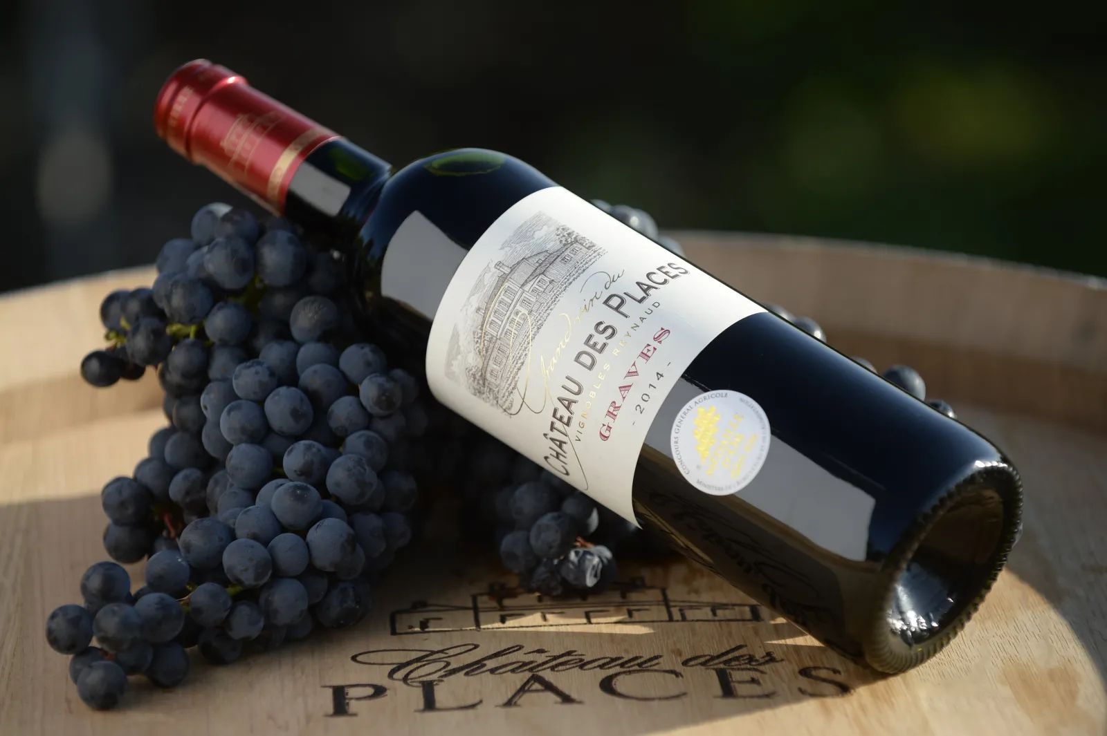 Red wine bottle Château des Places Bordeaux resting on wood with a bunch of black grapes, white label visible