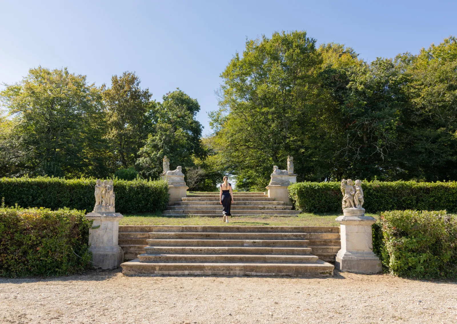 Monumental staircase of Château de Malle near Bordeaux, flanked by stone statues and trimmed hedges, with a woman climbing the steps under clear sky
