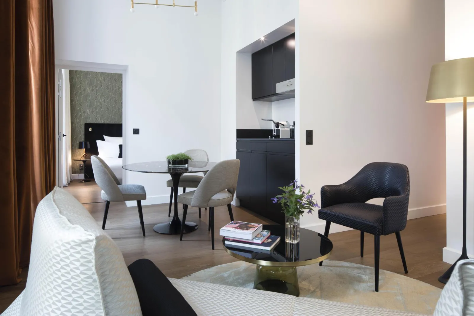 Elegant suite at Hotel Cardinal Bordeaux featuring modern living space, black kitchenette, contemporary gray and black furniture, and bed in background
