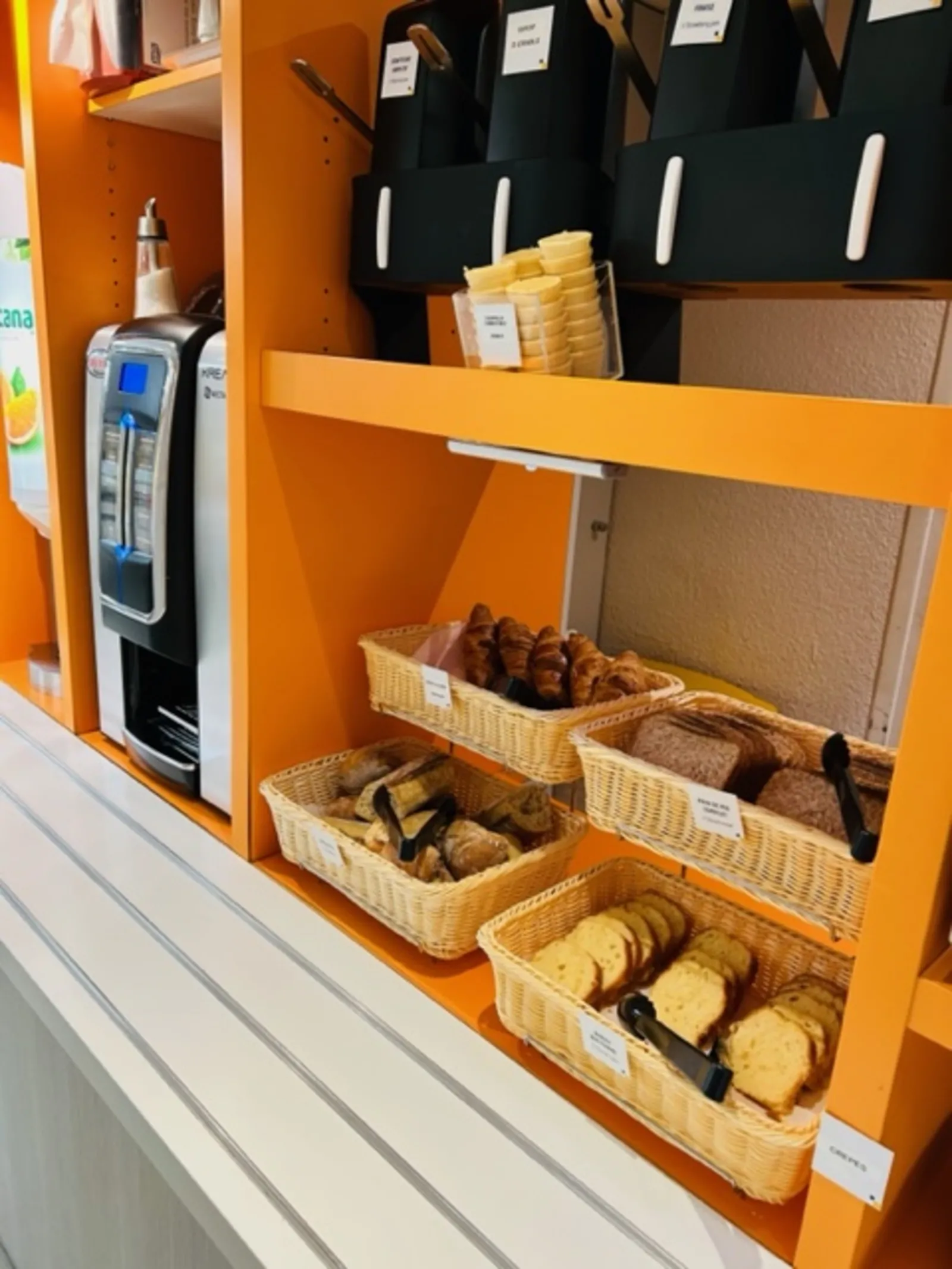 Breakfast display stand at Hôtel Première Classe Bordeaux South featuring wicker baskets with pastries, bread, and cookies, alongside a coffee machine and beverage dispenser.