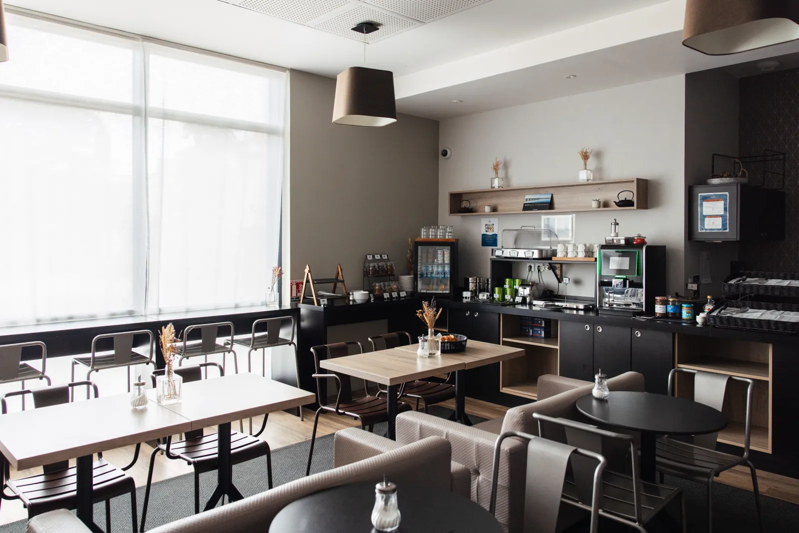 Modern common area in an aparthotel featuring a fully equipped kitchen, dining tables, designer chairs, large bright window, and contemporary black and natural wood decor.