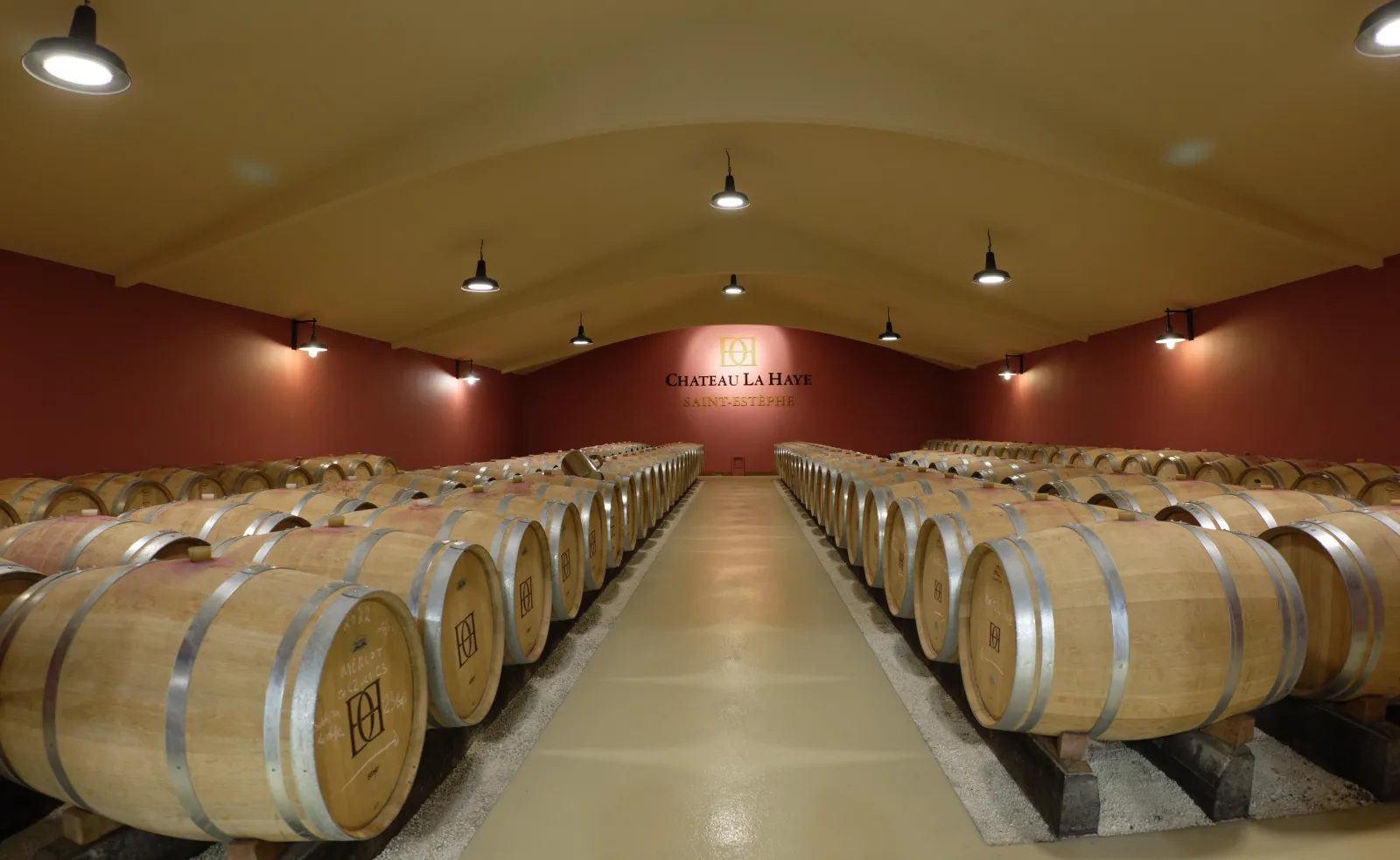 Aging cellar of Château La Haye in Bordeaux featuring rows of oak barrels symmetrically arranged under a vaulted ceiling with terracotta walls.