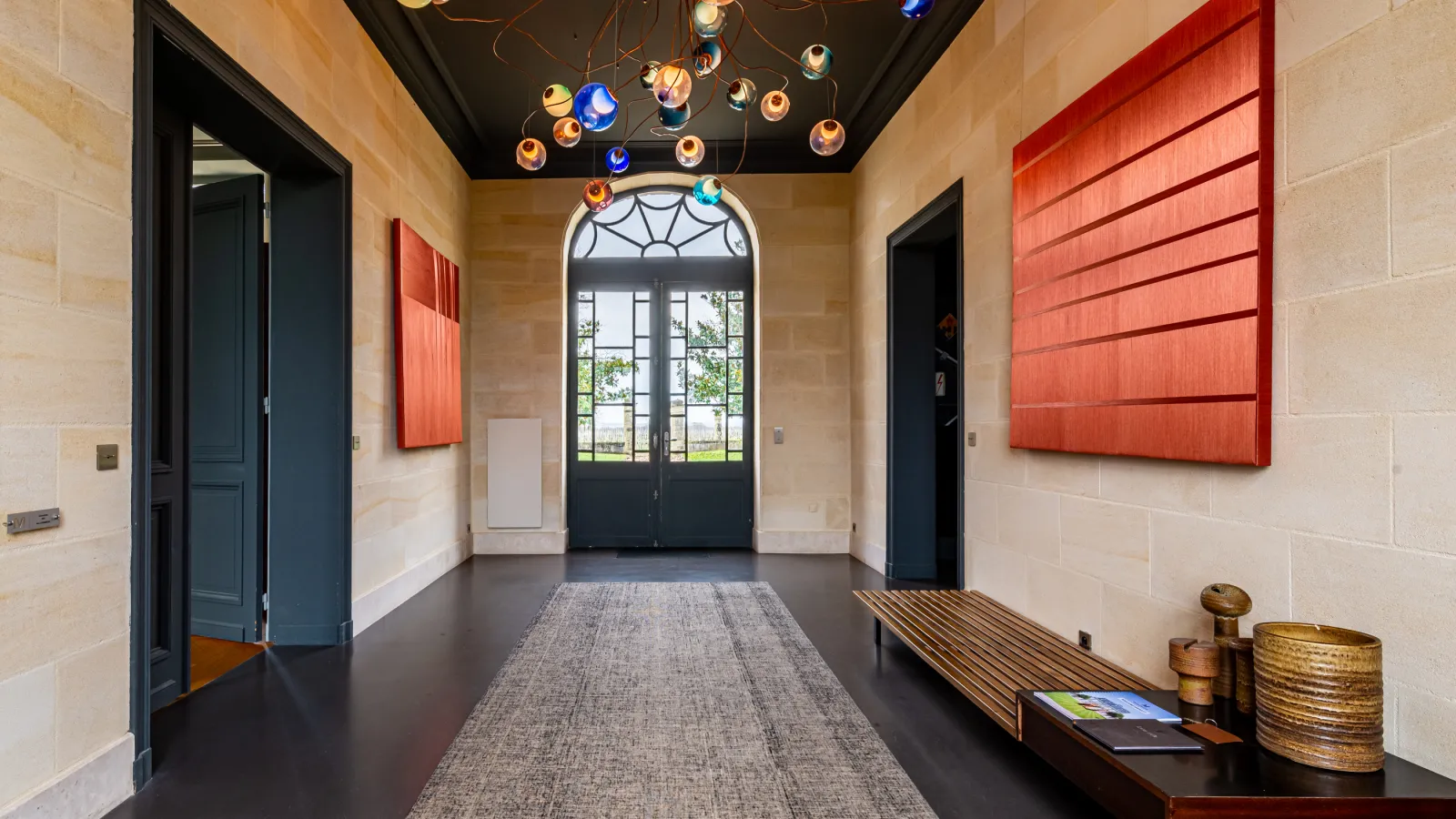 Elegant entrance hall of Château Malescasse in Bordeaux, featuring black doors, red panels, black ceiling with colorful pendant lights, and arched glass doors opening to gardens.