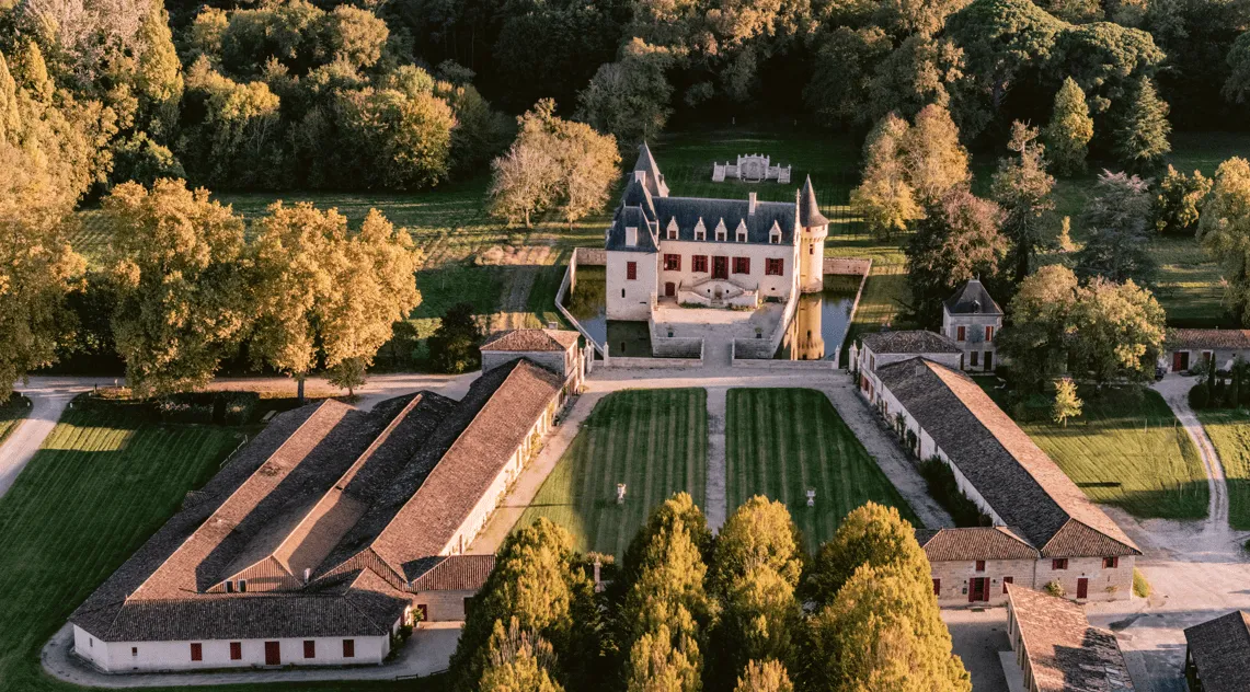 Aerial view of Château Olivier in Bordeaux, displaying the castle with its towers, surrounding vineyards, and wine production buildings, surrounded by autumn forest.