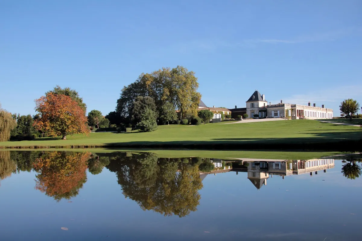 Château Paveil de Luze in Bordeaux, a classical white mansion surrounded by manicured grounds and autumn-colored trees, with its reflection mirrored in a tranquil pond in the foreground.