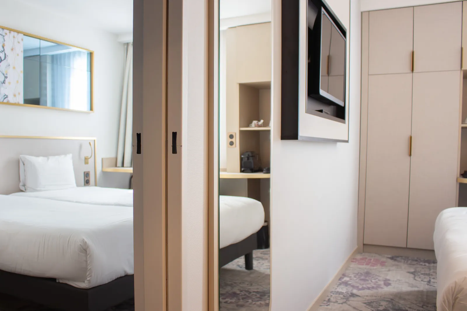 Modern hotel room featuring two beds, beige wardrobe, mirror, wall-mounted TV and ensuite bathroom at Brit Hôtel Bordeaux Aéroport Le Soretel