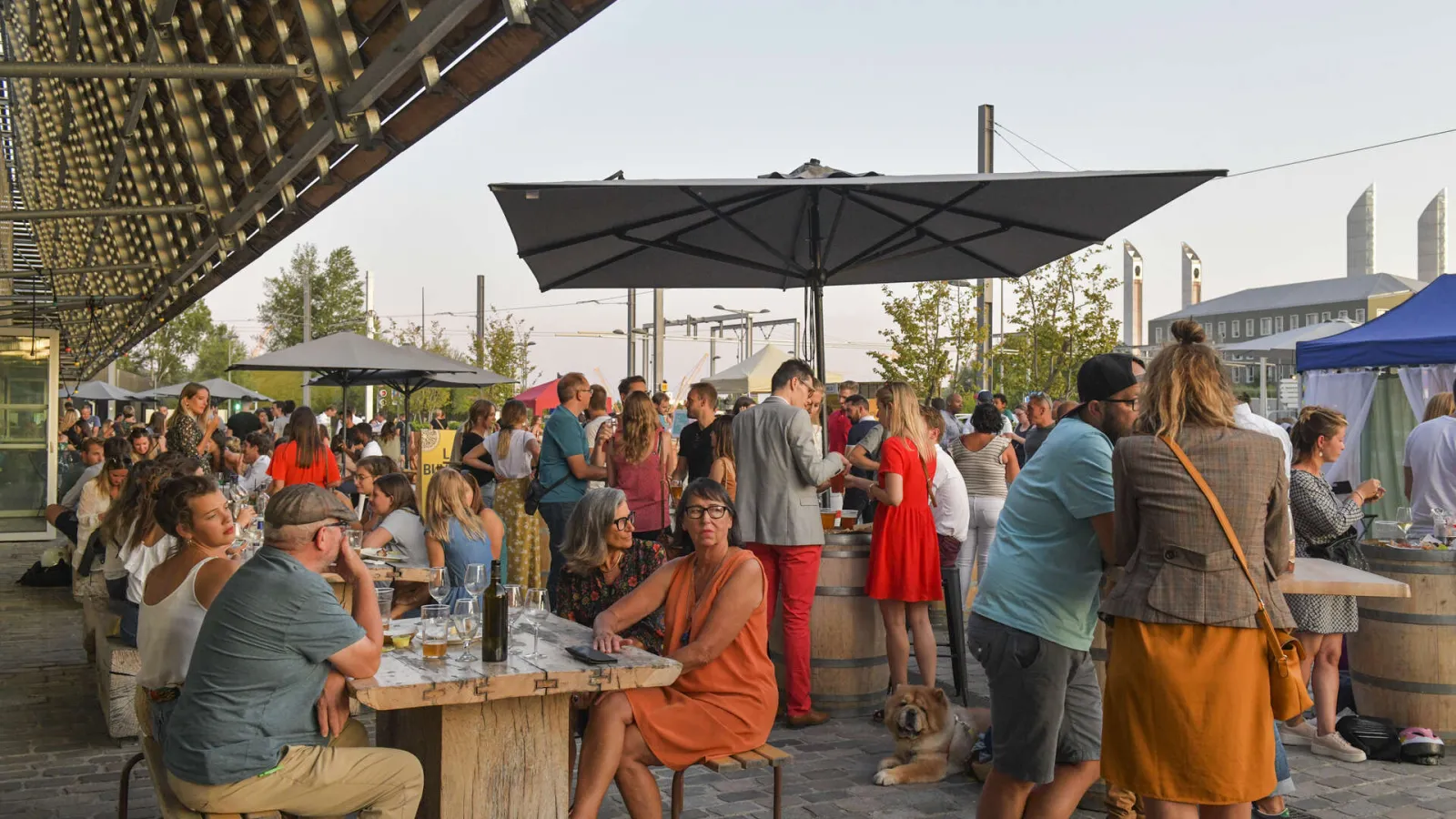 Crowd of visitors gathering at Halles de Bacalan in Bordeaux, enjoying a social moment under black umbrellas, with tables and drinks, modern architecture of the district visible in the background.