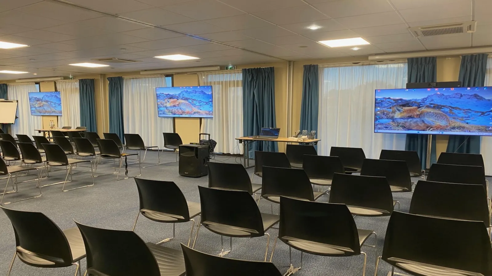 Modern conference room at Kyriad Prestige Bordeaux Ouest Mérignac hotel featuring black chairs, display screens showing mountain landscapes, and large windows with gray curtains.
