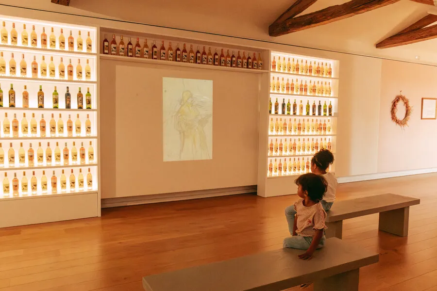 Two children sitting on a bench in an art gallery at Château Toulouse-Lautrec in Bordeaux, viewing illuminated displays of whisky bottles on the walls and artwork in the center.