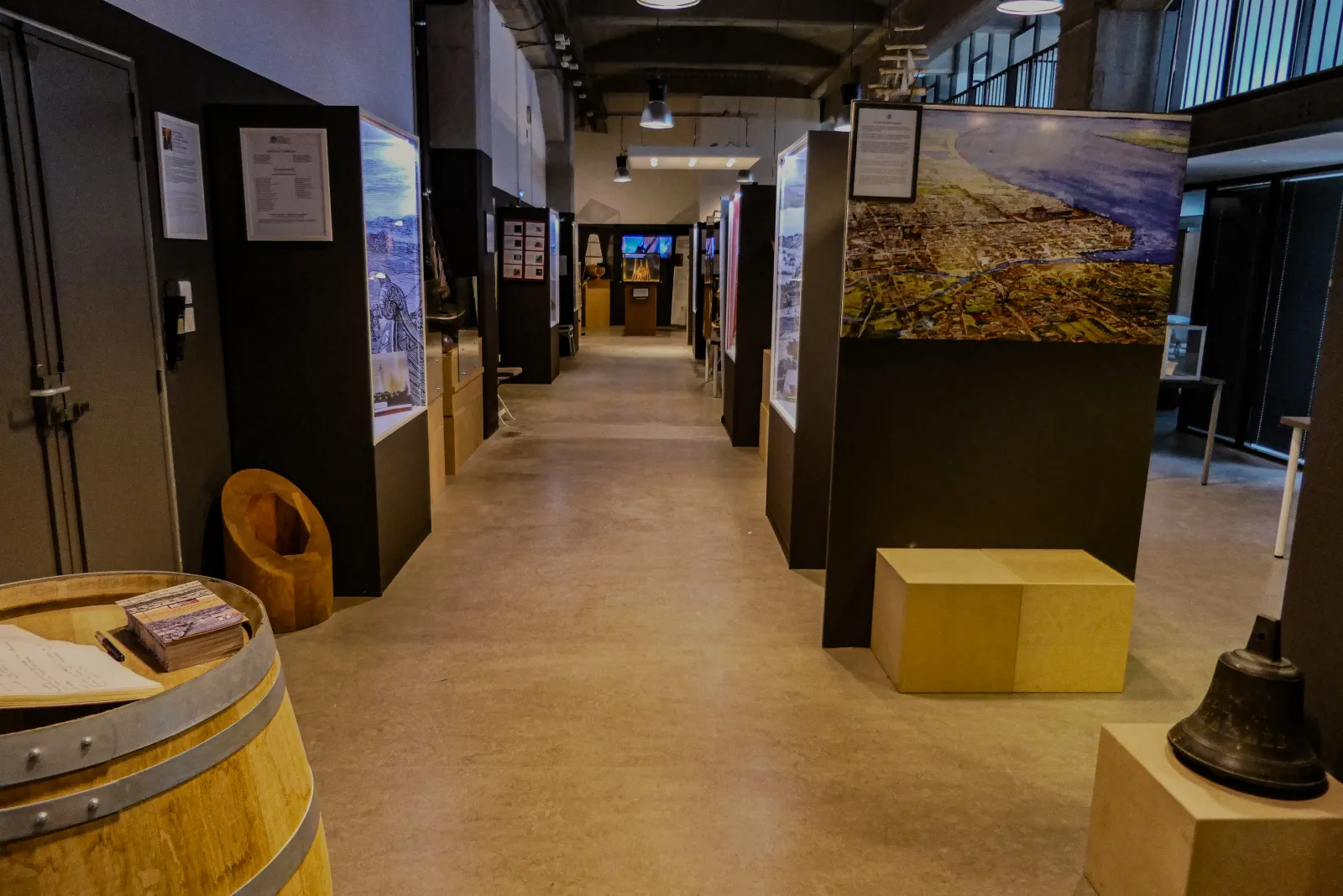 Interior of the Bordeaux Maritime History Museum displaying an exhibition gallery with informational panels, wooden barrels, display cases and historical maritime objects, lit by suspended lamps.