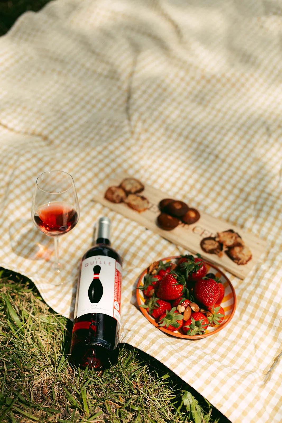 Picnic spread on a checkered white and beige blanket featuring a bottle of red wine from Château de Cérons, a glass of wine, fresh strawberries in a plate, walnuts and almonds, on green grass