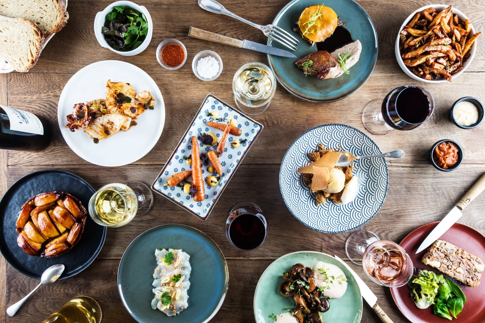 Tasting table with gourmet dishes, red and white wine glasses, roasted vegetables, meats and toasted bread arranged on colorful ceramic plates on wooden surface