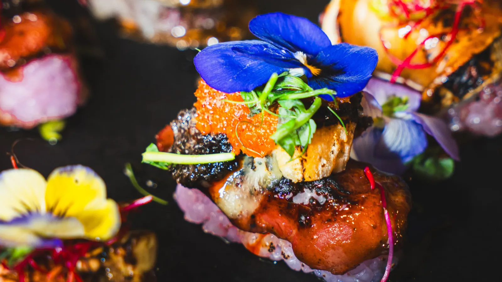 Gourmet burger topped with melted cheese, grilled vegetables and blue edible flowers, displayed on a dark background with colorful flowers
