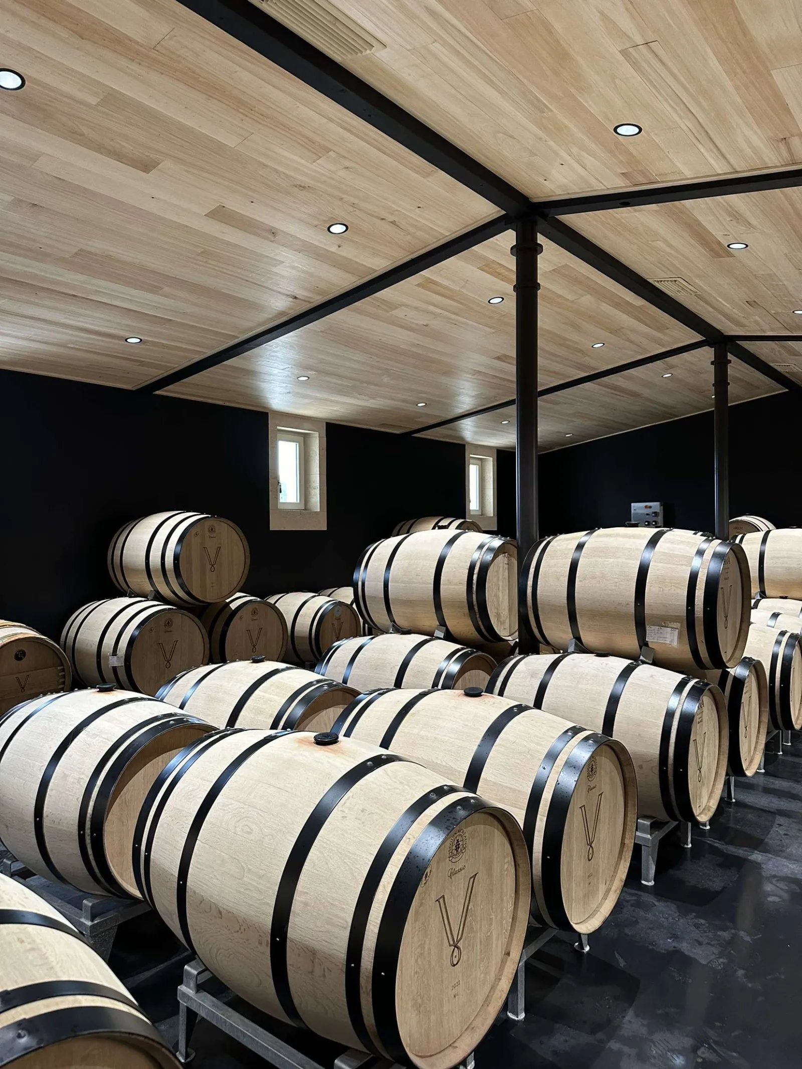 Wine cellar at Château Petit Val in Bordeaux featuring rows of wooden oak barrels arranged under a wooden ceiling with black metal framework