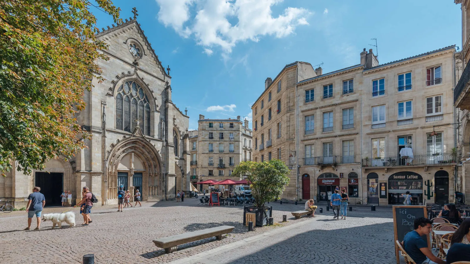Sunny square in Bordeaux featuring the Saint-Pierre Church with its ornate Gothic facade and clock, surrounded by Haussmann-style stone buildings, with visitors and café terraces.