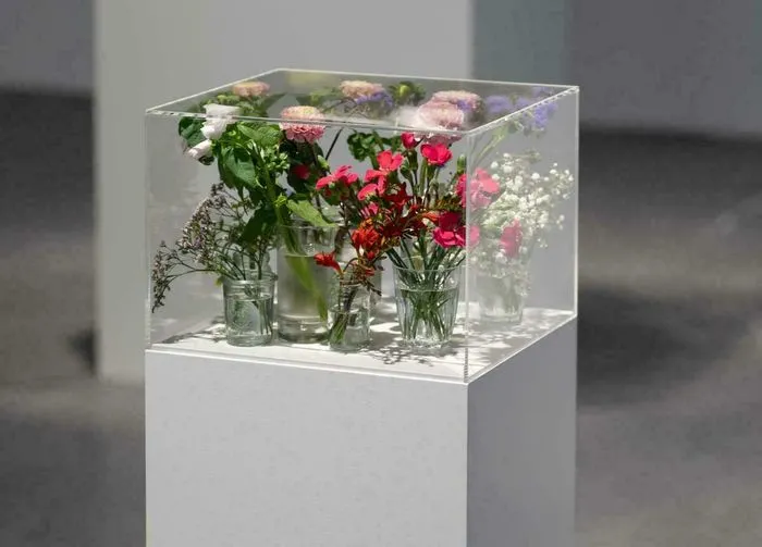 Artistic installation featuring a transparent glass display case containing multiple glass vases filled with fresh colorful flowers (roses, red and white carnations, baby's breath) on a white pedestal, Pollen exhibition at CAPC Bordeaux.