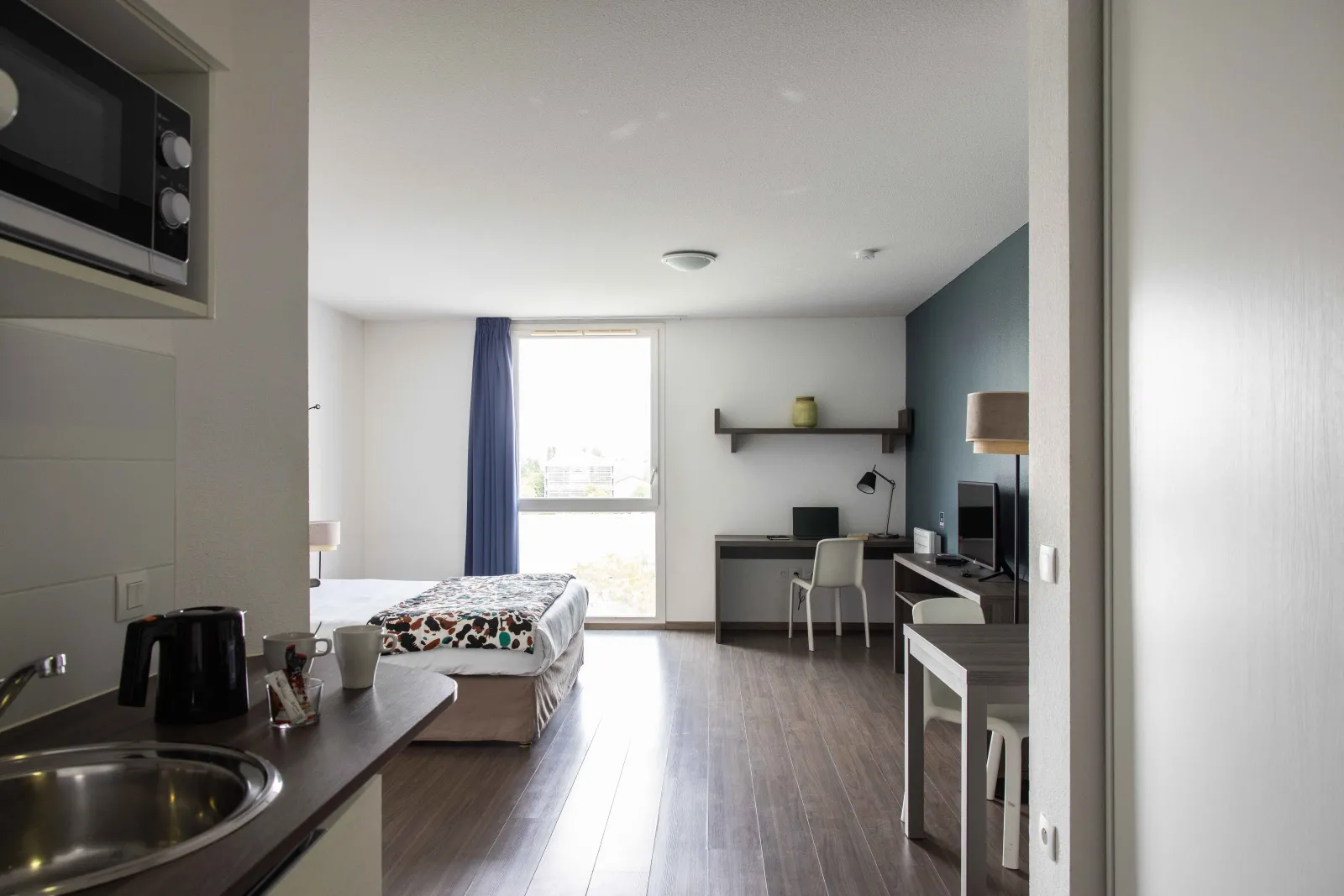 Modern studio apartment at Ténéo Aparthotel in Bordeaux-Maritime featuring a bed, equipped kitchenette, work area, large window with balcony and contemporary decor