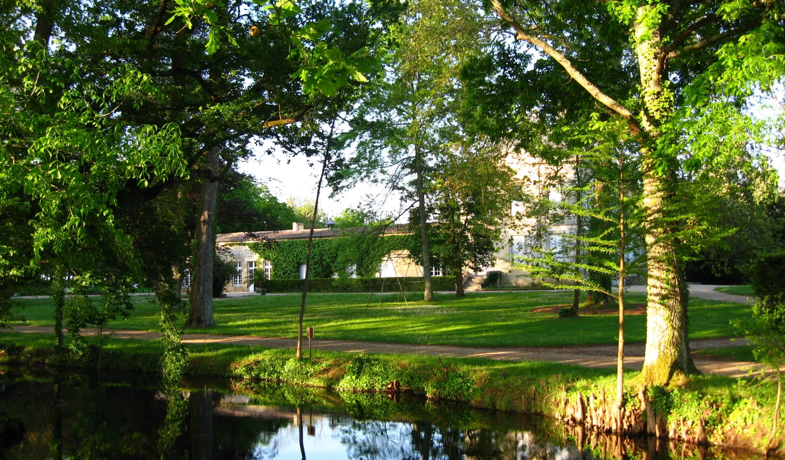 View of Château Siaurac in Bordeaux with a reflecting pond surrounded by majestic trees and lush green lawn, the main building visible in the background.