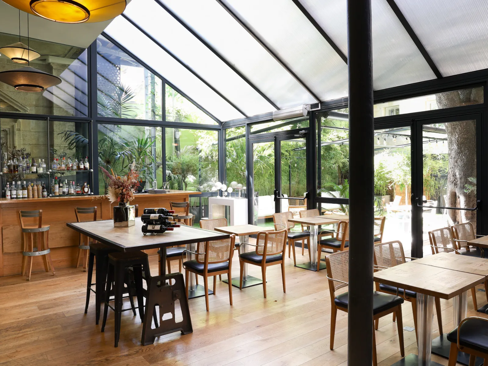 Upscale restaurant and bar space with glass ceiling, light wood furniture, elegant tables, and views of lush gardens in a Bordeaux boutique hotel.