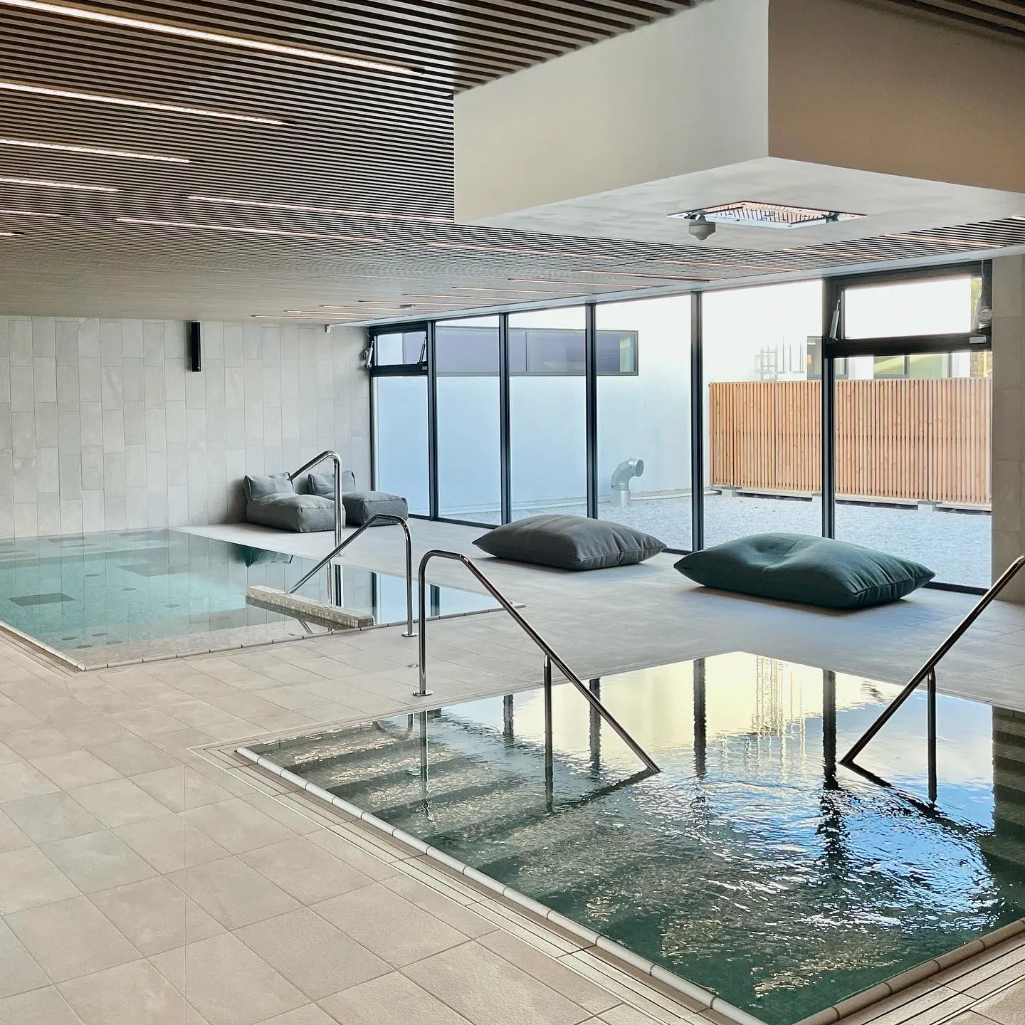 Modern aquatic wellness facility at UCPA Aqua Stadium in Mérignac near Bordeaux, featuring two relaxation pools, access stairs, lounging cushions, floor-to-ceiling windows, and covered outdoor terrace.