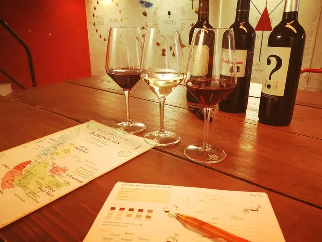 Bordeaux wine tasting session featuring three glasses filled with different wine colors, bottles in the background, and tasting notes sheets on a wooden table.