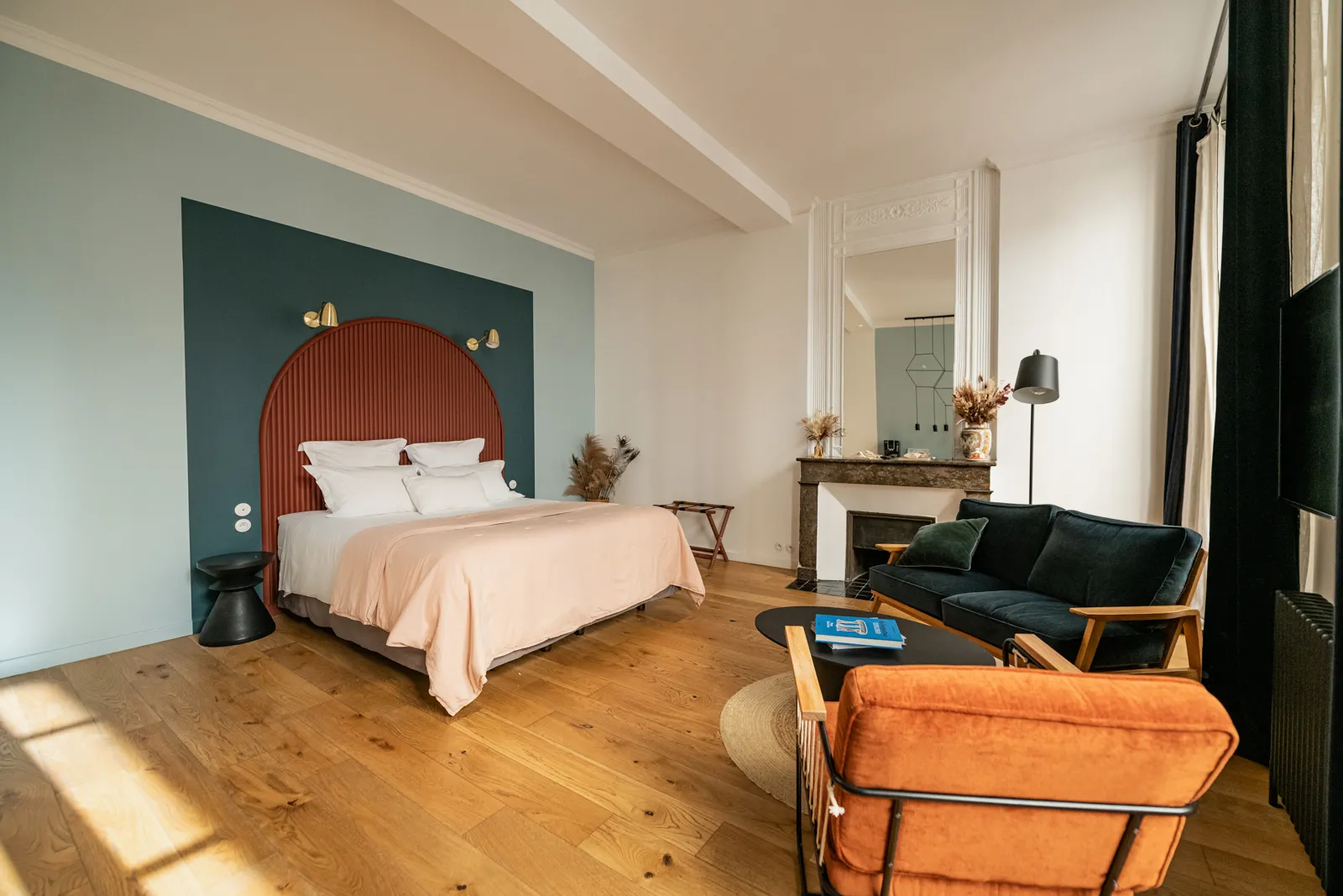 Spacious bedroom at La Maison Fernand in Bordeaux, featuring a king-size bed with red arched headboard, green and white painted walls, light wood flooring, sitting area with black sofa and orange armchair, decorated fireplace and large windows.