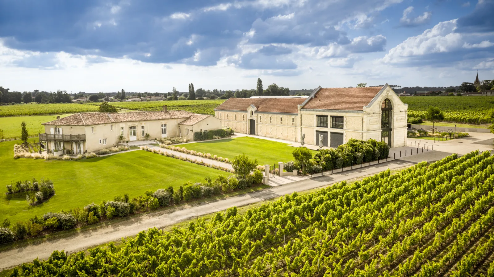 Château La Garde in Bordeaux featuring white stone buildings surrounded by lush vineyards and manicured lawns under a partly cloudy sky