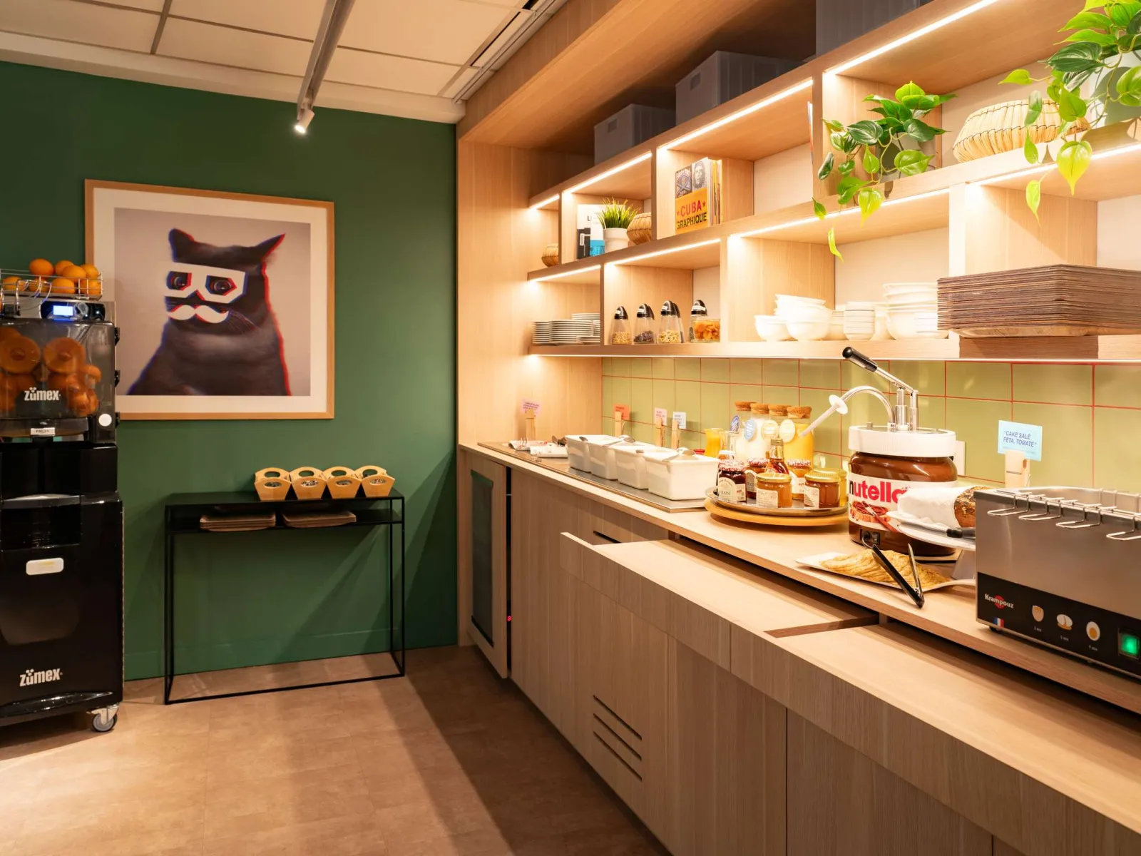Modern and bright kitchen at Ibis Styles Bordeaux Centre Gare hotel featuring green walls, illuminated shelves, white countertops, professional equipment, and a stylized cat wall decoration.