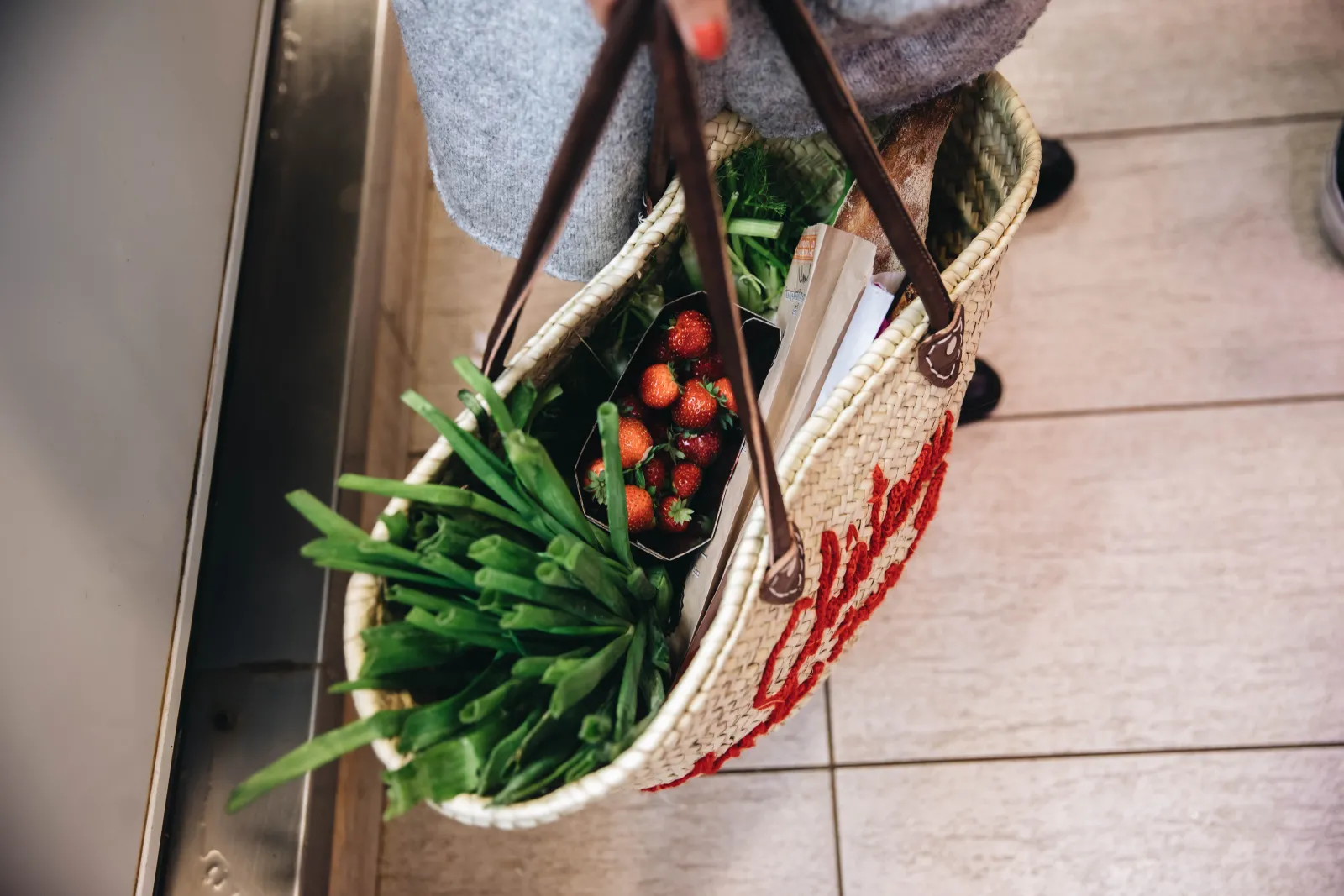 Person holding a wicker basket filled with fresh strawberries and green leeks, wearing gray clothing and a brown shoulder bag, standing on a tiled floor.
