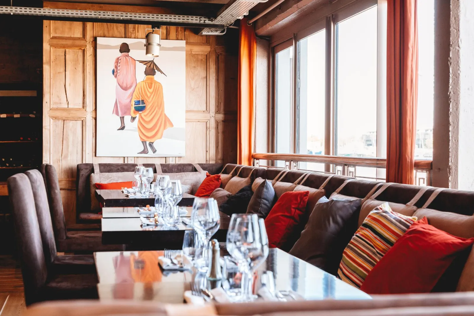 Elegant interior of Bordeaux's Maritime Café featuring brown leather banquettes, red cushions, dining tables with glassware, floor-to-ceiling windows overlooking the city, and artwork depicting figures in colorful robes.