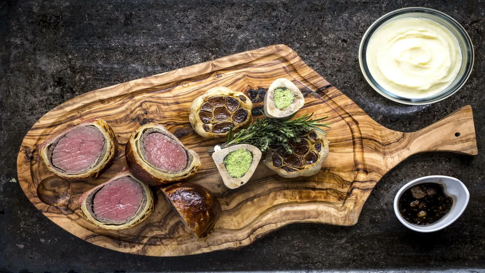 Gourmet tasting platter featuring individual meat pies, seafood topped with green paste, accompanied by creamy sauce and dark reduction on olive wood board.
