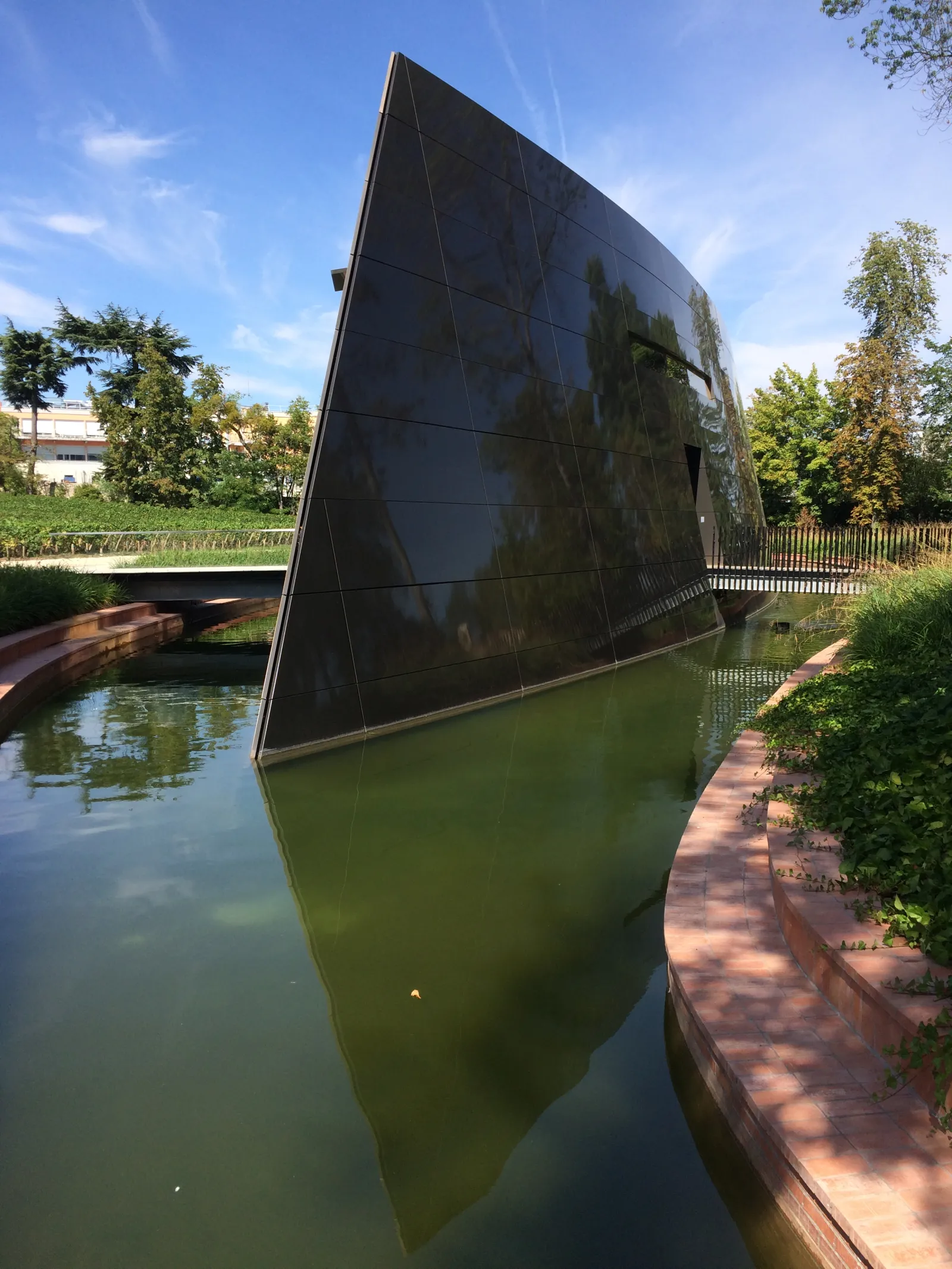 A modern black tilted architectural structure overlooking a canal in Bordeaux, with reflections in the water and surrounding vegetation