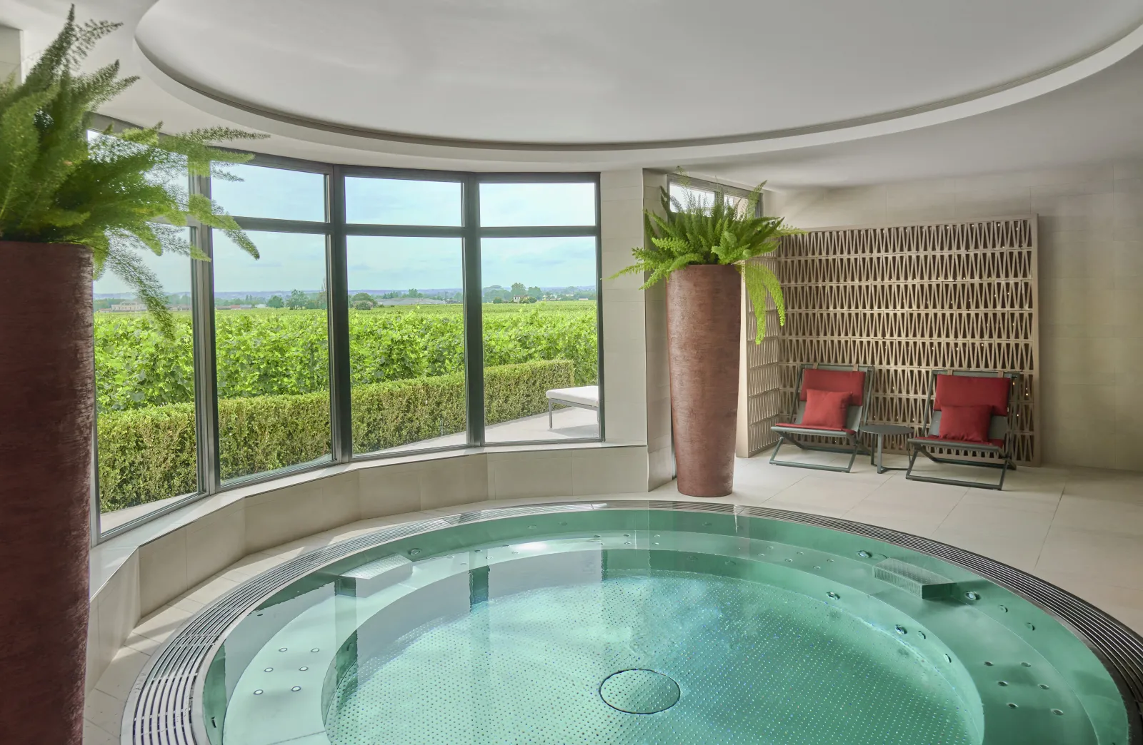Indoor spa with turquoise circular hot tub, panoramic windows overlooking Bordeaux vineyards, potted green plants, red lounge chairs, and curved ceiling.